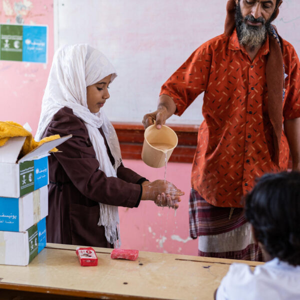 Standing at the front of a classroom, a schoolgirl washes her hands in water that a man pours from a jug.