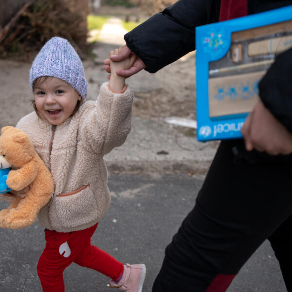 A three-year-old holding a teddy bear is on a walk with a guardian outside.