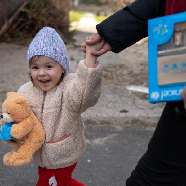 A three-year-old holding a teddy bear is on a walk with a guardian outside.