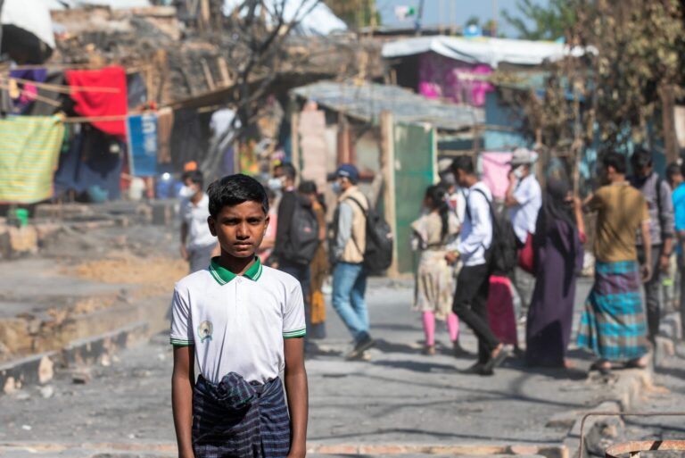 A 13-year-old boy stands facing the camera while Rohingya refugees pass through the refugee camp in the background.