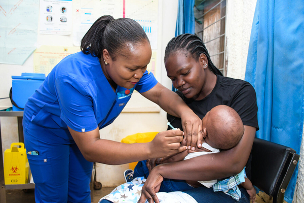 A 6-month-old baby is held by his seated mother, while being vaccinated by a health worker in a hospital.